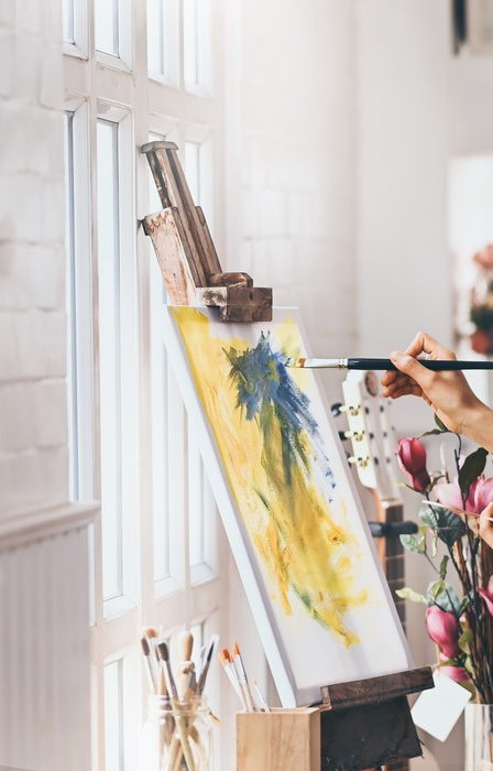 A woman creates an abstract blue and yellow painting on a canvas. She sits in a clean white room with natural lighting.