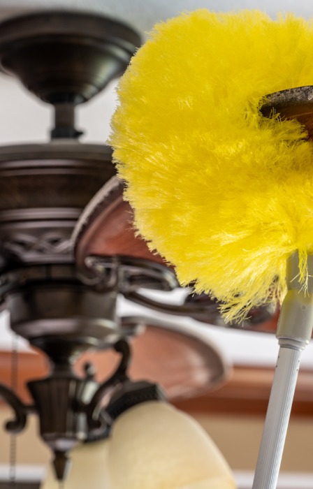 A close-up of a yellow duster wrapped around a ceiling fan blade. The fan blade is dusty and being cleaned.