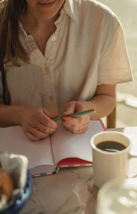 A close-up of someone sitting at a table with an open notebook in front of them. There is also a cup of black coffee.