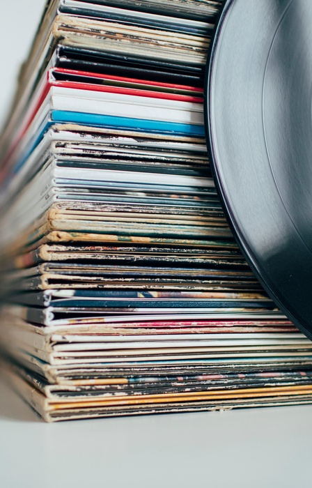 A stack of vinyl records in their original album jackets sit in a pile on a white surface. An exposed vinyl leans against it.