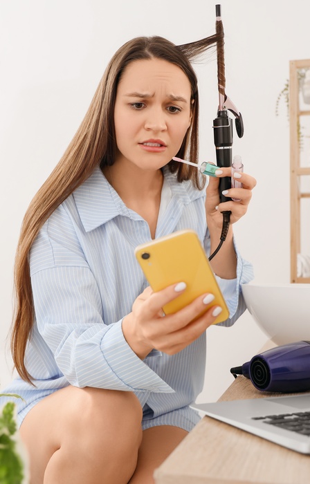 A woman checks her phone while curling her hair next to a laptop and hair dryer on a bathroom counter.