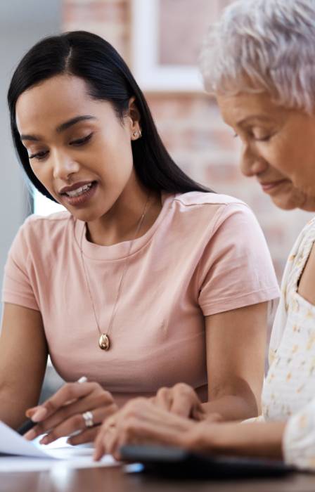 A woman reviews paperwork at a table with an older woman seated beside her as they look over the documents together.