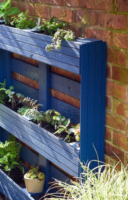 A vertical herb garden made out of a blue painted wooden pallet is leaning against a brick wall outdoors.