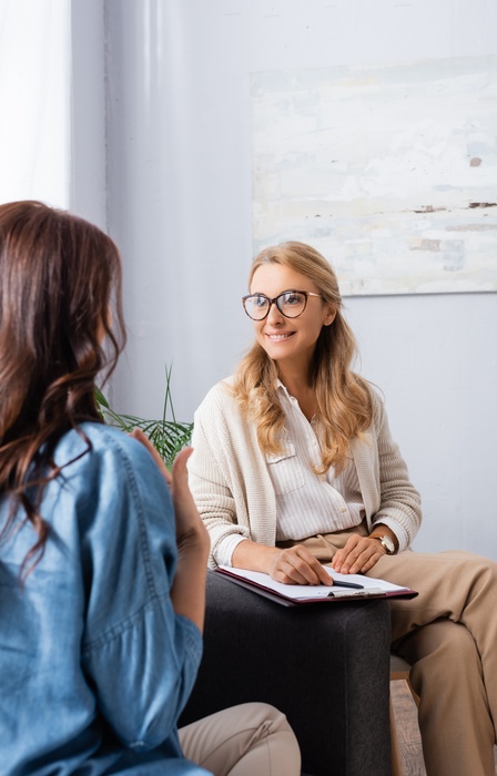 A woman seen from behind sitting on a couch across from another woman holding a clipboard during a conversation.