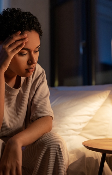 A woman sitting in her bed at night, holding her head with one hand while looking at the clock next to her table lamp.