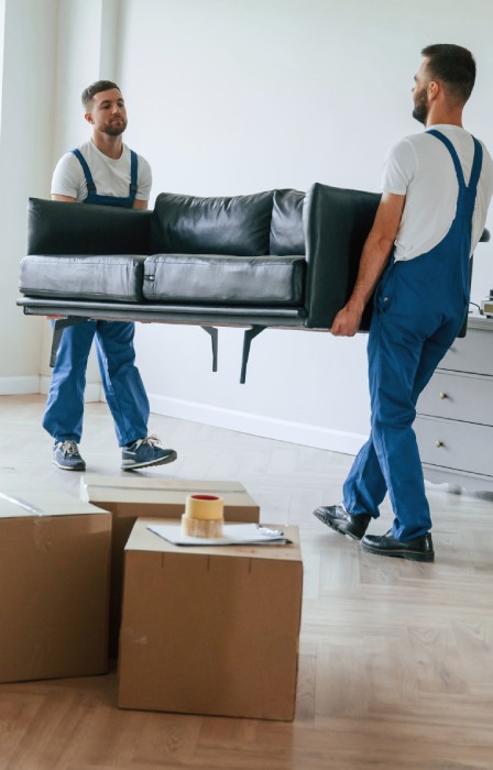Two men wear denim overalls and white T-shirts as they carry a couch out of a home. There are boxes stacked up.