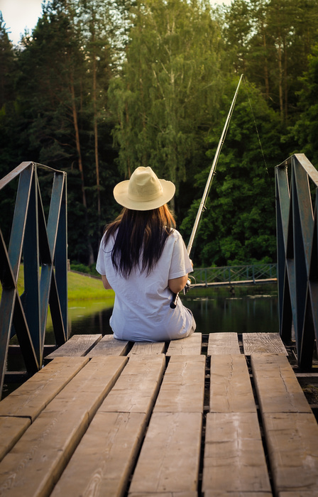 A woman in a white t-shirt and a light hat sits on the edge of a dock, fishing on a calm lake or pond with her hair down.