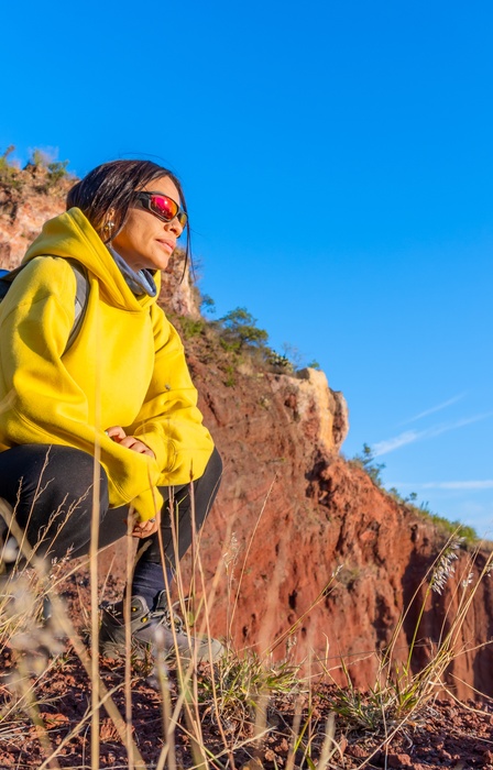 A hiker is wearing a yellow hoodie and backpack while squatting on a rocky hillside, overlooking a mountain landscape.