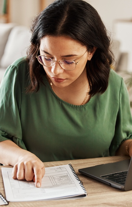 A woman sits at a desk in her home, comparing the financial information in her notebook to that on her computer.