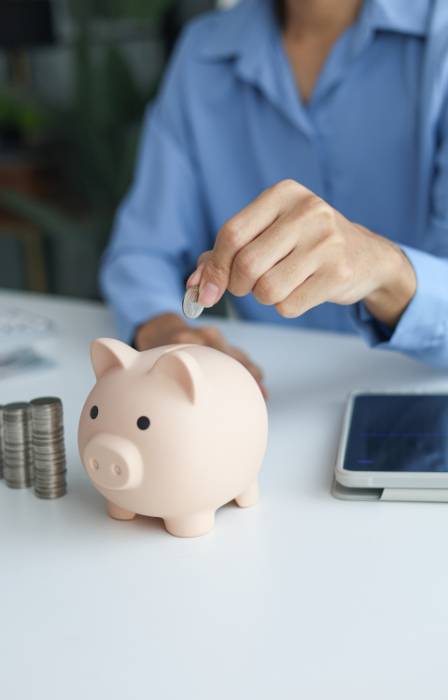 A person wearing a blue blouse is dropping a silver coin into the top opening of a pink piggy bank.