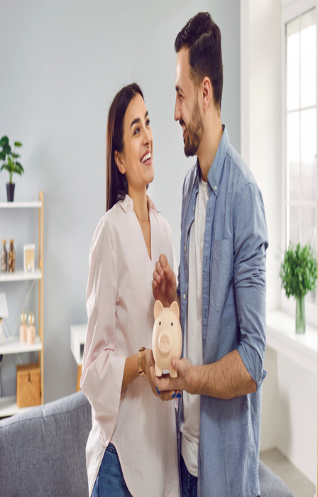 A couple smiles as they look at each other. They both hold a piggy bank, and the man goes to put money inside.