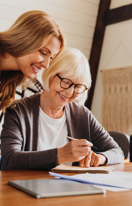 An older woman sitting down at the table to sign documents with her adult daughter over her shoulder to review them.