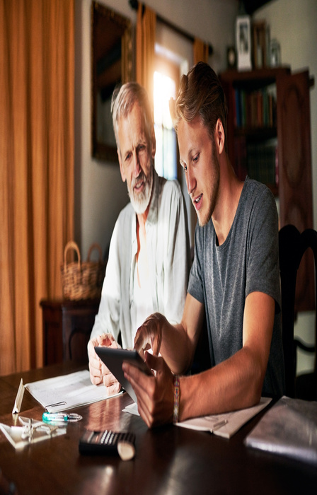 A father and son sitting at a table look at a tablet and discuss finances. The sun shines through a window behind them.