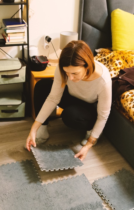 a woman crouching on a bedroom floor while assembling gray interlocking foam mats near a bed and storage shelves.
