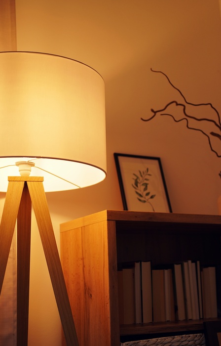 A warm lamp with a wide shade and a tripod base standing beside a bookshelf and a curtain.